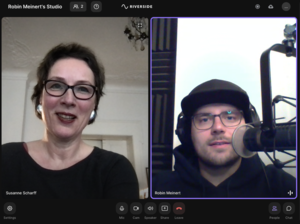 Podcast-Interview mit Susanne Scharff und Robin Meißner, beide im Gespräch vor Mikrofonen. Susanne trägt eine schwarze Bluse und hat eine freundliche Mimik, Robin trägt eine schwarze Mütze und Brille.