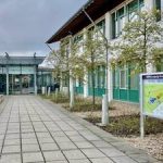 Außenansicht des BBW Leipzig mit gepflastertem Weg, Bäumen und Informationsschild.