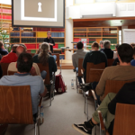 Eine Gruppe von Menschen sitzt in einem Raum und hört einem Vortrag zu. Im Hintergrund ist eine Präsentation mit einem Schlüsselbild zu sehen. Die Teilnehmer sind in einem modernen Bibliotheksumfeld.