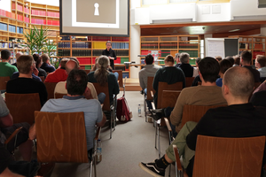 Eine Gruppe von Menschen sitzt in einem Raum und hört einem Vortrag zu. Im Hintergrund ist eine Präsentation mit einem Schlüsselbild zu sehen. Die Teilnehmer sind in einem modernen Bibliotheksumfeld.