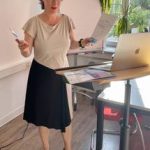 Referentin steht an einem Tisch mit Laptop, hält Unterlagen in der Hand, trägt ein beiges Oberteil und einen schwarzen Rock.