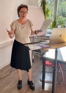 Referentin steht an einem Tisch mit Laptop, hält Unterlagen in der Hand, trägt ein beiges Oberteil und einen schwarzen Rock.